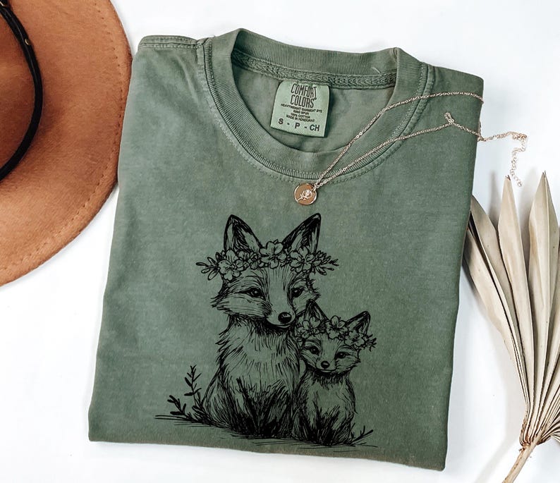 May include: An olive green Comfort Colors t-shirt featuring a black illustration of two foxes wearing floral crowns. A gold necklace with a small pendant rests on the shirt. The shirt is folded, with a brown hat visible in the background.