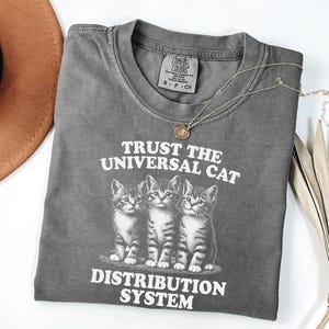 May include: A gray t-shirt with the text "TRUST THE UNIVERSAL CAT DISTRIBUTION SYSTEM" and an illustration of three kittens. A gold necklace with a pendant is draped across the shirt.