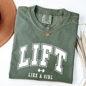 Pode incluir: Camiseta verde-oliva com a palavra "LIFT" em letras brancas grandes, um gr&aacute;fico de halteres e a frase "LIKE A GIRL". Um colar dourado e um chap&eacute;u marrom tamb&eacute;m s&atilde;o vis&iacute;veis.