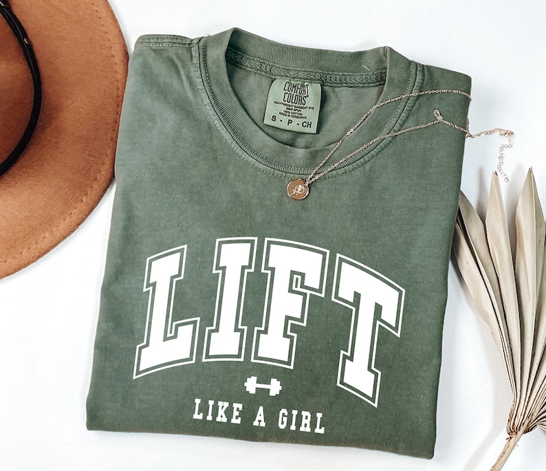 Pode incluir: Camiseta verde-oliva com a palavra "LIFT" em letras brancas grandes, um gr&aacute;fico de halteres e a frase "LIKE A GIRL". Um colar dourado e um chap&eacute;u marrom tamb&eacute;m s&atilde;o vis&iacute;veis.
