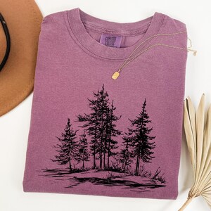 May include: A mauve-colored t-shirt with a crew neck and a black graphic of several pine trees. A gold necklace with a small pendant is draped across the shirt. The shirt has a Comfort Colors tag.