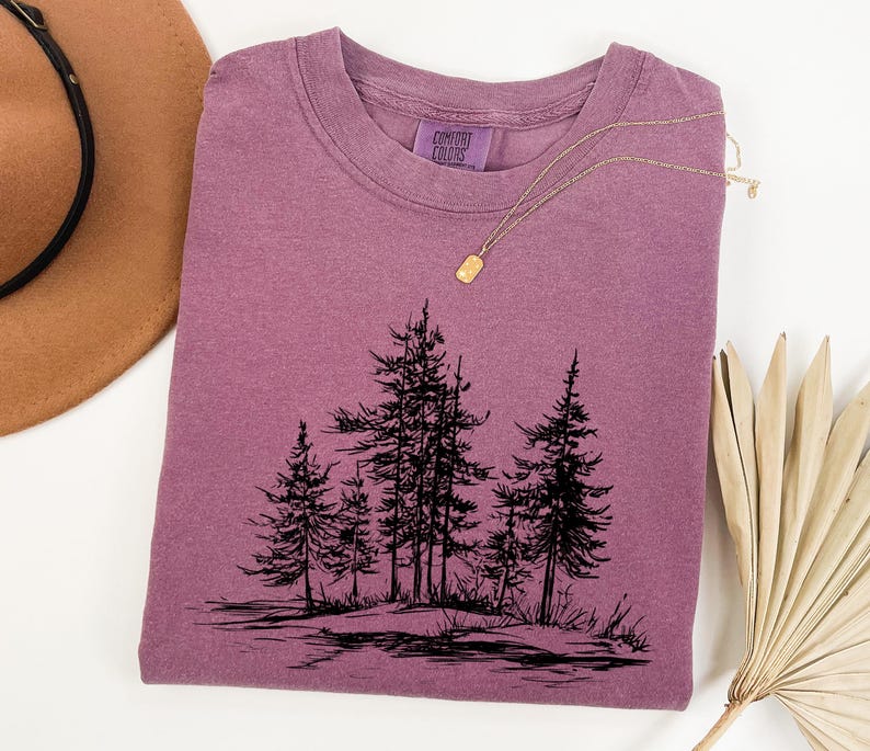 May include: A mauve-colored t-shirt with a crew neck and a black graphic of several pine trees. A gold necklace with a small pendant is draped across the shirt. The shirt has a Comfort Colors tag.