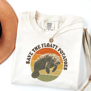 May include: A cream-colored short-sleeve t-shirt featuring a graphic of a manatee centered within a circular sunset design in orange, yellow, and green. The text "SAVE THE FLOATY POTATOES" is printed in a dark brown arched font around the graphic.