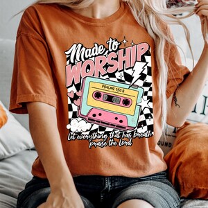 Puede incluir: Una camiseta de color &oacute;xido con un gr&aacute;fico de cinta de casete retro y el texto "Made to Worship" en rosa y blanco. Tambi&eacute;n se incluye el texto "Psalms 150:6". El gr&aacute;fico incluye un patr&oacute;n de cuadros y un rayo.