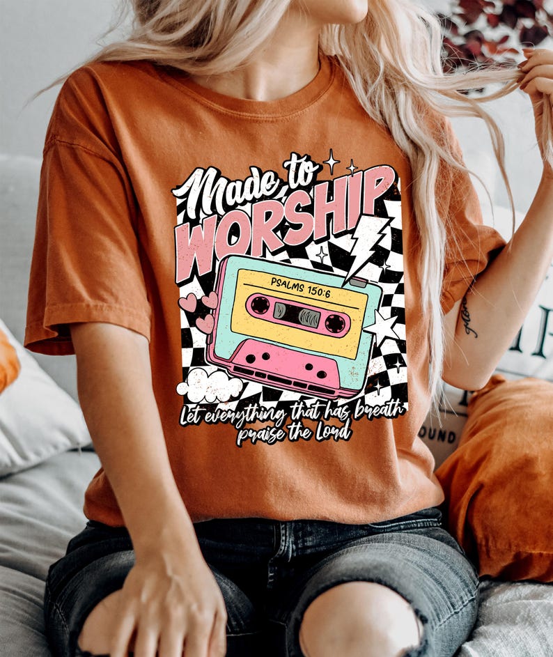 Puede incluir: Una camiseta de color &oacute;xido con un gr&aacute;fico de cinta de casete retro y el texto "Made to Worship" en rosa y blanco. Tambi&eacute;n se incluye el texto "Psalms 150:6". El gr&aacute;fico incluye un patr&oacute;n de cuadros y un rayo.