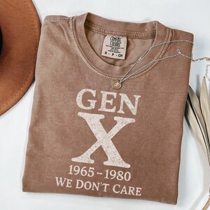 May include: A brown t-shirt with the text "GEN X 1965-1980 WE DON'T CARE" in white. A silver necklace with a pendant is draped across the shirt. The shirt is folded, and a brown hat is visible in the upper left corner.