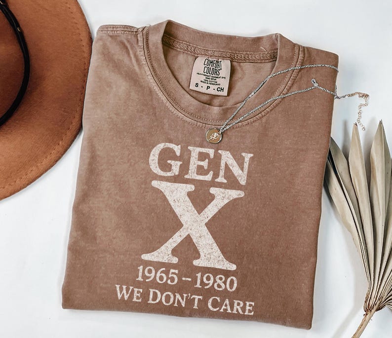 May include: A brown t-shirt with the text "GEN X 1965-1980 WE DON'T CARE" in white. A silver necklace with a pendant is draped across the shirt. The shirt is folded, and a brown hat is visible in the upper left corner.