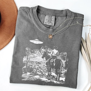 May include: Dark gray Comfort Colors t-shirt featuring a graphic of a cowboy on horseback with a UFO in the sky. The shirt has a crew neck and short sleeves. A gold necklace with a pendant is draped on the shirt.