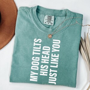以下が含まれることがあります： 「MY DOG TILTS HIS HEAD JUST LIKE YOU」の白い文字が入った、シーフォームグリーンのTシャツ。小さなペンダントが付いたゴールドのネックレスがシャツの上にあります。茶色の帽子と乾燥した植物が背景にあります。