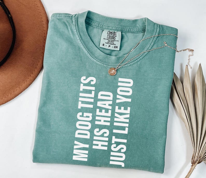 以下が含まれることがあります： 「MY DOG TILTS HIS HEAD JUST LIKE YOU」の白い文字が入った、シーフォームグリーンのTシャツ。小さなペンダントが付いたゴールドのネックレスがシャツの上にあります。茶色の帽子と乾燥した植物が背景にあります。