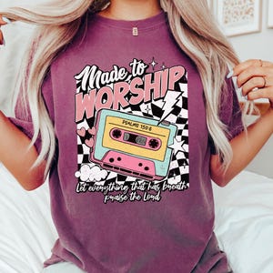 Puede incluir: Una camiseta morada con un dise&ntilde;o gr&aacute;fico retro que presenta una cinta de casete, estrellas y el texto "Made to Worship" con el vers&iacute;culo b&iacute;blico "Salmos 150:6" y la frase "Que todo lo que tiene aliento alabe al Se&ntilde;or".