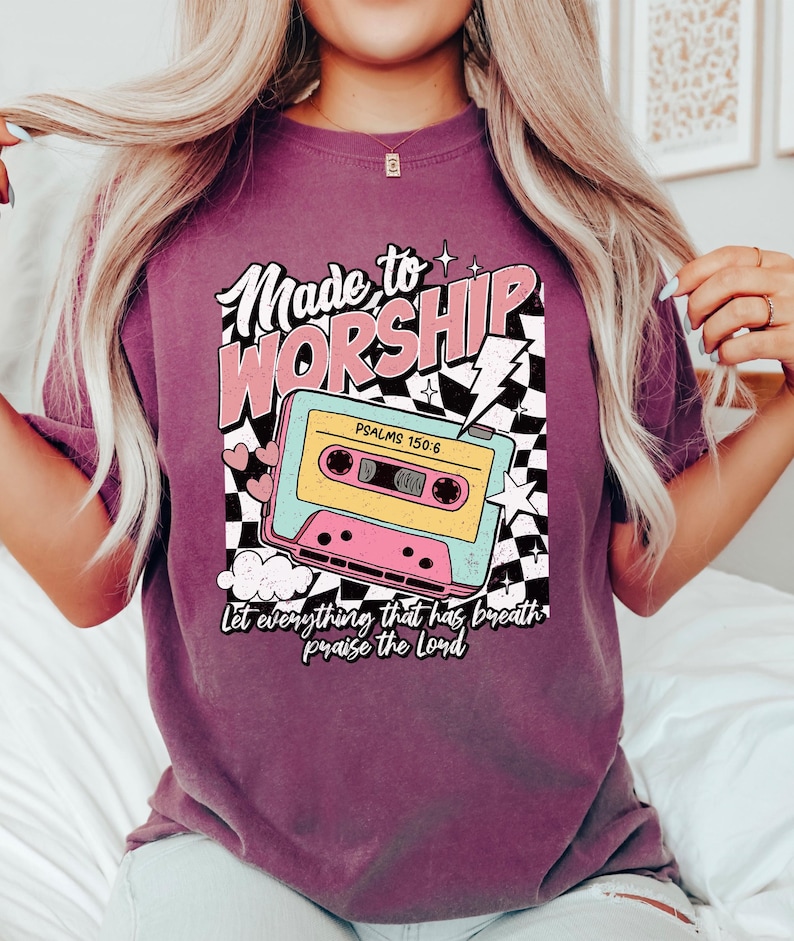 Puede incluir: Una camiseta morada con un dise&ntilde;o gr&aacute;fico retro que presenta una cinta de casete, estrellas y el texto "Made to Worship" con el vers&iacute;culo b&iacute;blico "Salmos 150:6" y la frase "Que todo lo que tiene aliento alabe al Se&ntilde;or".