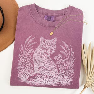 May include: A mauve Comfort Colors t-shirt featuring a white illustration of a fox surrounded by flowers and foliage. A gold necklace rests on the shirt. A brown hat is visible in the upper left corner.