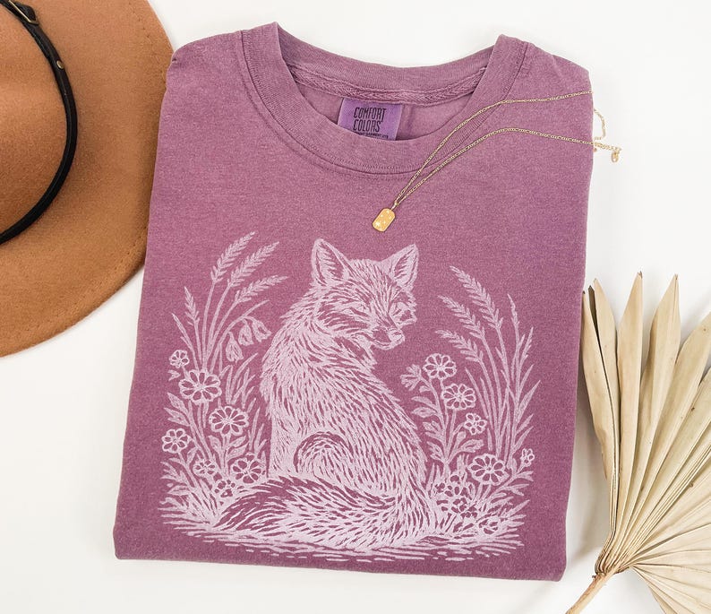 May include: A mauve Comfort Colors t-shirt featuring a white illustration of a fox surrounded by flowers and foliage. A gold necklace rests on the shirt. A brown hat is visible in the upper left corner.