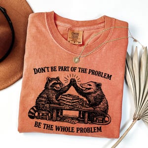 May include: Orange Comfort Colors t-shirt with the text "DON'T BE PART OF THE PROBLEM BE THE WHOLE PROBLEM." The graphic shows a raccoon and opossum high-fiving over a table with pizza. A gold necklace and a brown hat are also in the image.