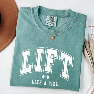 Pode incluir: Uma camiseta verde-azulado com o texto "LIFT" em letras brancas grandes, um pequeno gr&aacute;fico de halteres e as palavras "LIKE A GIRL" abaixo. Um colar de prata com um pingente redondo repousa sobre a camisa. Um chap&eacute;u marrom e uma planta seca est&atilde;o no fundo.