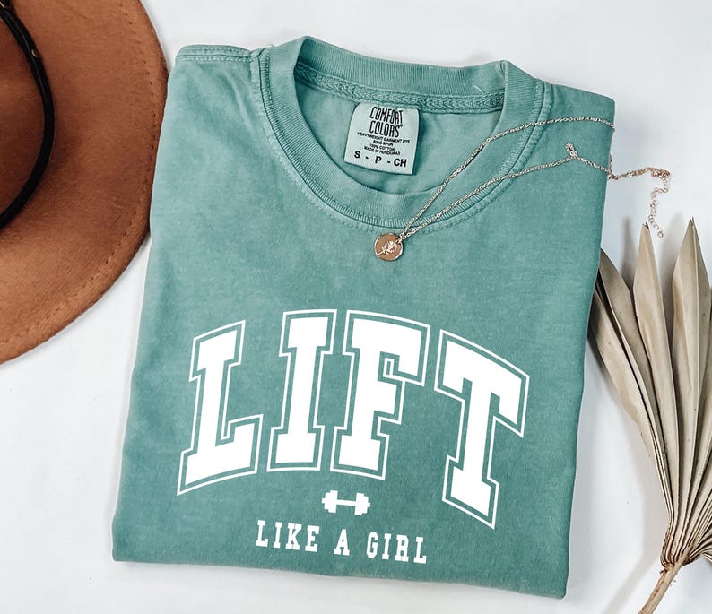 Pode incluir: Uma camiseta verde-azulado com o texto "LIFT" em letras brancas grandes, um pequeno gr&aacute;fico de halteres e as palavras "LIKE A GIRL" abaixo. Um colar de prata com um pingente redondo repousa sobre a camisa. Um chap&eacute;u marrom e uma planta seca est&atilde;o no fundo.