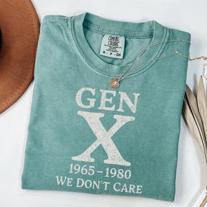 May include: A teal Comfort Colors t-shirt with the text "GEN X 1965-1980 WE DON'T CARE" in white. A silver necklace with a circular pendant rests on the shirt. A brown hat and dried plant are in the background.