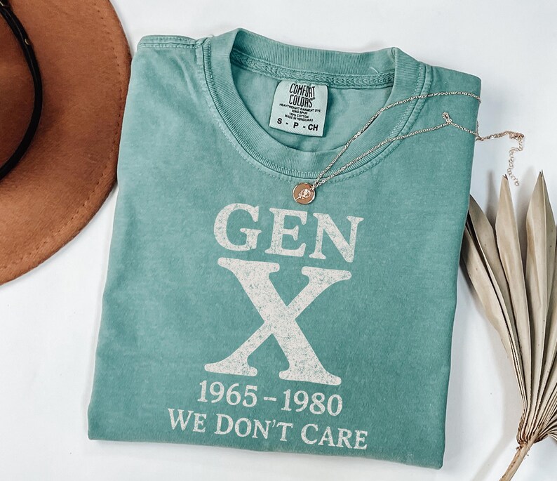 May include: A teal Comfort Colors t-shirt with the text "GEN X 1965-1980 WE DON'T CARE" in white. A silver necklace with a circular pendant rests on the shirt. A brown hat and dried plant are in the background.