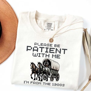 May include: A beige t-shirt with the text "Please be patient with me I'm from the 1990s" in black pixelated font. A vintage-style illustration of a horse-drawn wagon is below the text.