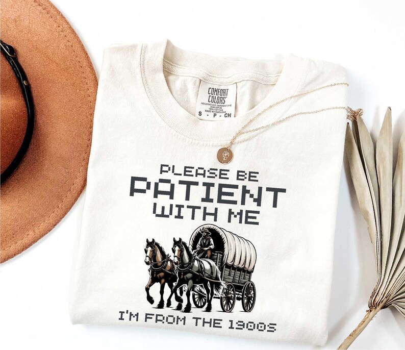 May include: A beige t-shirt with the text "Please be patient with me I'm from the 1990s" in black pixelated font. A vintage-style illustration of a horse-drawn wagon is below the text.