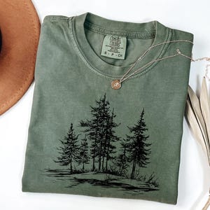 May include: An olive green t-shirt with a black graphic of a forest scene. The shirt has a crew neck and short sleeves. A gold necklace with a pendant rests on the shirt. The shirt is folded on a white surface.