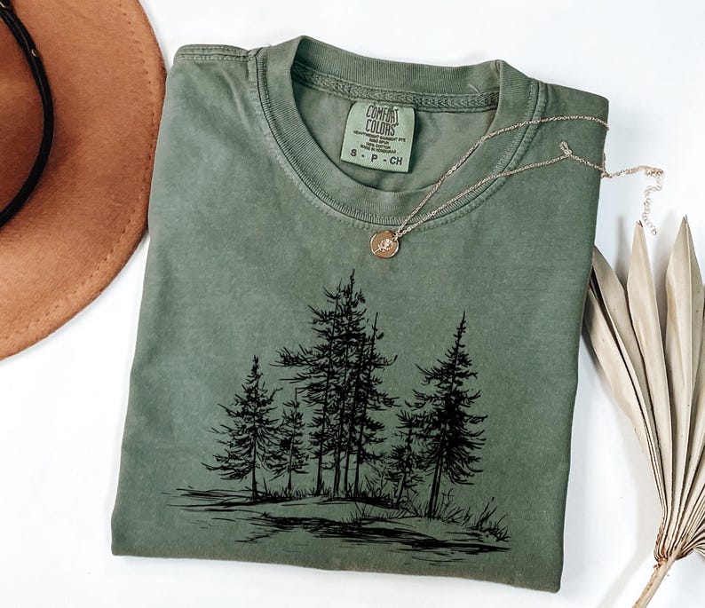 May include: An olive green t-shirt with a black graphic of a forest scene. The shirt has a crew neck and short sleeves. A gold necklace with a pendant rests on the shirt. The shirt is folded on a white surface.