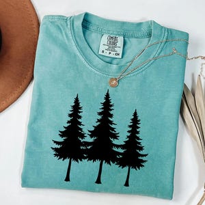 May include: A teal Comfort Colors t-shirt featuring a black graphic of three pine trees. The shirt has a crew neck and short sleeves. A gold necklace with a pendant is draped across the shirt.