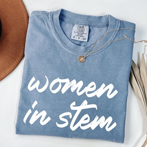May include: A blue t-shirt with the words "Women in STEM" in white script. The shirt is folded and has a gold necklace draped across it. A brown hat and dried plant are in the background.