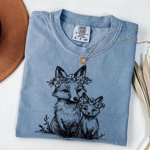 May include: A blue Comfort Colors t-shirt featuring a black illustration of two foxes wearing floral crowns. A gold necklace with a pendant rests on the shirt. The shirt is folded on a white surface.