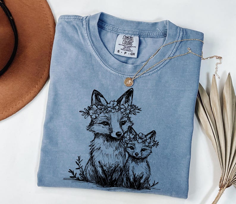 May include: A blue Comfort Colors t-shirt featuring a black illustration of two foxes wearing floral crowns. A gold necklace with a pendant rests on the shirt. The shirt is folded on a white surface.
