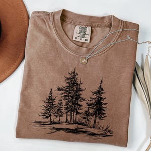 May include: A brown Comfort Colors t-shirt with a crew neck and short sleeves. The shirt features a black graphic of pine trees and a silver necklace with a pendant. The shirt is size S-P-CH.