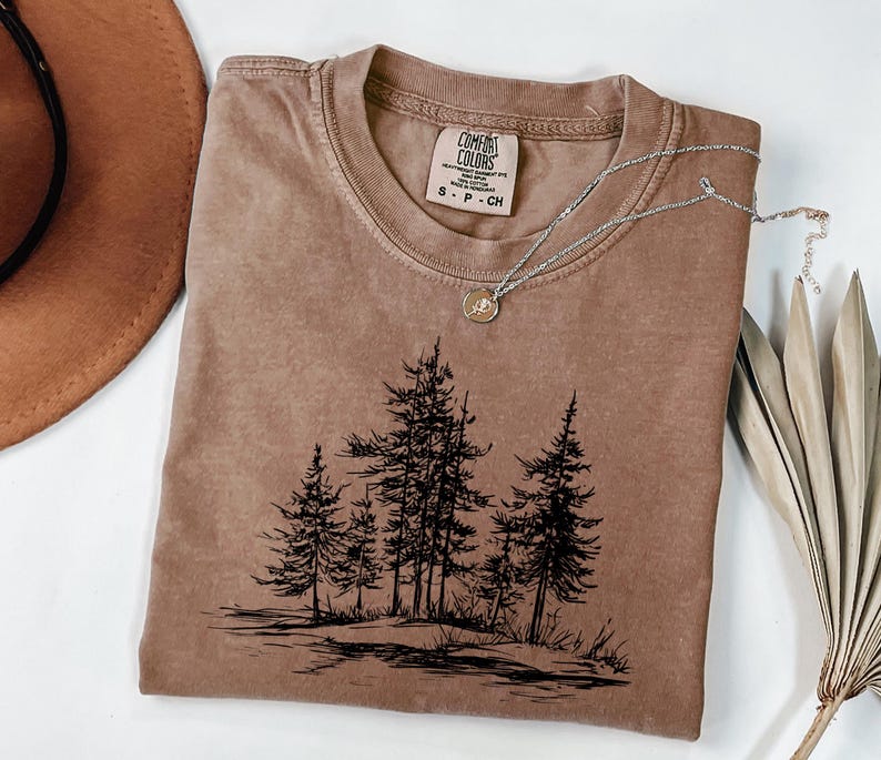 May include: A brown Comfort Colors t-shirt with a crew neck and short sleeves. The shirt features a black graphic of pine trees and a silver necklace with a pendant. The shirt is size S-P-CH.