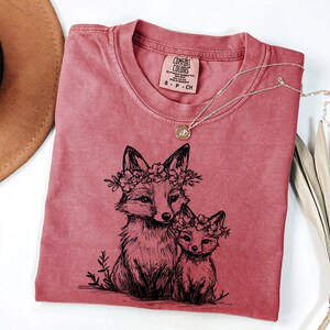 May include: A dusty rose Comfort Colors t-shirt featuring a black illustration of two foxes wearing floral crowns. A delicate silver necklace with a small pendant rests on the shirt. The shirt has a crew neck and a small tag.