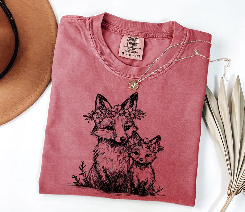 May include: A dusty rose Comfort Colors t-shirt featuring a black illustration of two foxes wearing floral crowns. A delicate silver necklace with a small pendant rests on the shirt. The shirt has a crew neck and a small tag.