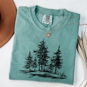 May include: A teal Comfort Colors t-shirt with a black graphic of pine trees. A silver necklace with a circular pendant rests on the shirt. The shirt is folded, and a brown hat is visible in the background.