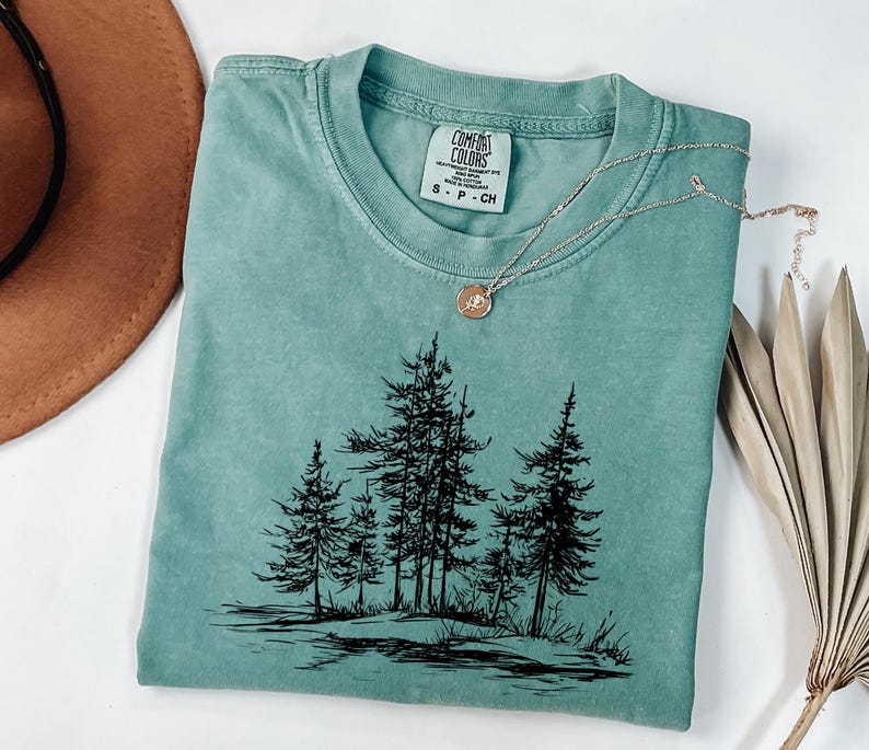 May include: A teal Comfort Colors t-shirt with a black graphic of pine trees. A silver necklace with a circular pendant rests on the shirt. The shirt is folded, and a brown hat is visible in the background.
