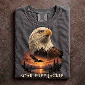 May include: A charcoal gray short-sleeve t-shirt featuring a large graphic of a bald eagle head and a smaller eagle in flight over a lake, mountains, and sunset. The text SOAR FREE JACKIE is printed at the bottom in a light beige serif font.