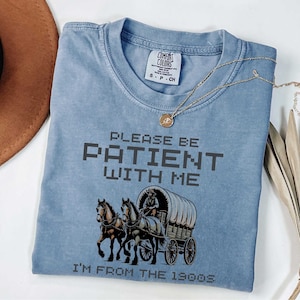 May include: A light blue t-shirt with a vintage-style graphic of a horse-drawn wagon and the text "Please be patient with me I'm from the 1900s".