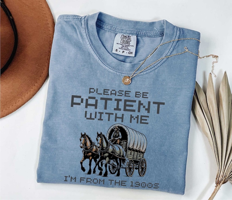 May include: A light blue t-shirt with a vintage-style graphic of a horse-drawn wagon and the text "Please be patient with me I'm from the 1900s".
