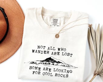 May include: Cream-colored Comfort Colors t-shirt with the text "NOT ALL WHO WANDER ARE LOST SOME ARE LOOKING FOR COOL ROCKS" in black. A gold necklace and a brown hat are also visible.