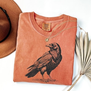 May include: A coral-colored crew neck t-shirt featuring a detailed black illustration of a raven. The shirt has a vintage wash appearance and a small tag at the neckline. A gold necklace with a pendant is draped across the shirt.