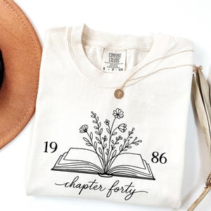 May include: Cream-colored t-shirt with a black graphic of an open book with flowers sprouting from it. The text "1986 chapter forty" is below the book. A gold necklace and a brown hat are also in the image.