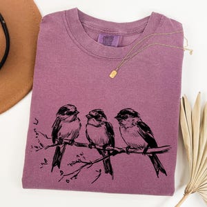 May include: A mauve-colored t-shirt with a crew neck featuring a black illustration of three birds perched on a branch. A gold necklace and a brown hat are also in the image. The shirt has a Comfort Colors tag.
