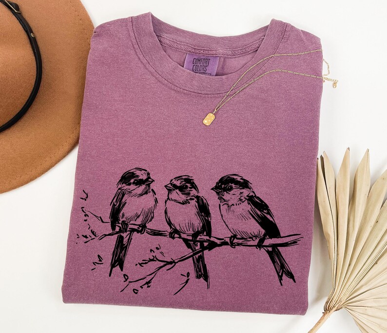 May include: A mauve-colored t-shirt with a crew neck featuring a black illustration of three birds perched on a branch. A gold necklace and a brown hat are also in the image. The shirt has a Comfort Colors tag.