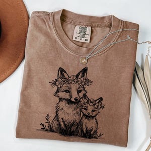 May include: A brown Comfort Colors t-shirt featuring a black illustration of two foxes wearing floral crowns. A silver necklace with a small pendant rests on the shirt. The shirt is folded, with a brown hat visible in the background.