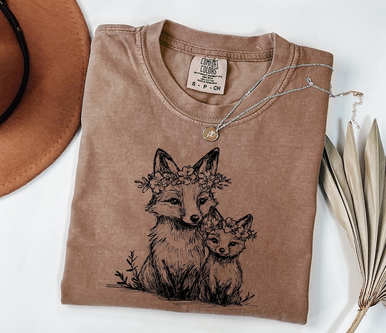 May include: A brown Comfort Colors t-shirt featuring a black illustration of two foxes wearing floral crowns. A silver necklace with a small pendant rests on the shirt. The shirt is folded, with a brown hat visible in the background.