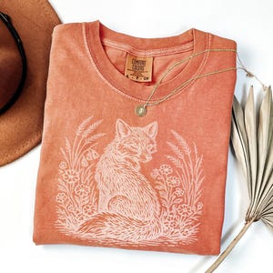 May include: A coral-colored t-shirt with a round neckline and a white graphic of a fox surrounded by flowers and wheat. A gold necklace with a pendant rests on the shirt. A brown hat is visible in the upper left corner.