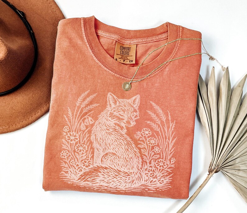 May include: A coral-colored t-shirt with a round neckline and a white graphic of a fox surrounded by flowers and wheat. A gold necklace with a pendant rests on the shirt. A brown hat is visible in the upper left corner.