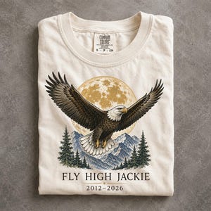 May include: A cream-colored t-shirt featuring a graphic of a bald eagle in flight before a large full moon and mountain range with pine trees. The text reads FLY HIGH JACKIE 2012-2026. The shirt has a Comfort Colors brand label at the neck.